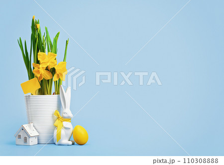 Yellow Easter eggs in a nest and blooming daffodils in a white pot and Easter bunny with tiny house on blue background. Happy Easter wide web banner 110308888