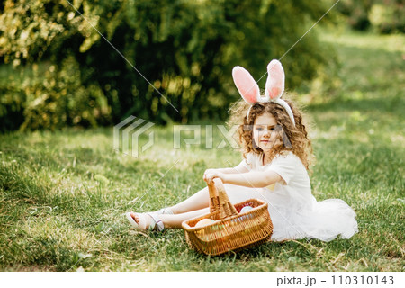 Easter egg hunt. Girl child Wearing Bunny Ears Running To Pick Up Egg In Garden. Easter tradition. Baby with basket full of colorful eggs. 110310143