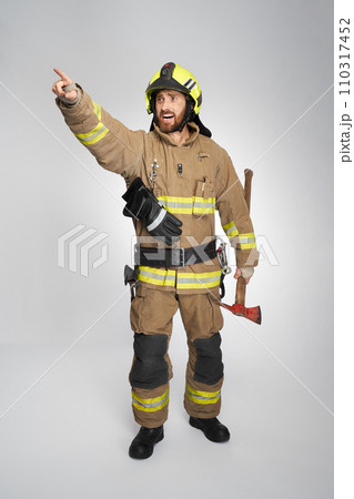Concerned firefighter in uniform shouting, while pointing away indoors. Front view of excited bearded fireman showing direction, while carrying fire axe, on gray background. Concept of work, rescue. Concerned firefighter in uniform shouting, while pointing away indoors. Front view of excited bearded fireman showing direction, while carrying fire axe, on gray background. Concept of work, rescue. 110317452