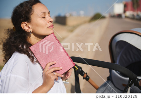 Multiracial woman enjoys reading a book outdoor, relaxing on the park with her cute baby sleeping in the baby pram 110320129