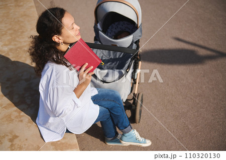 Young mother enjoys reading a book outdoor, relaxing on the park with her cute baby sleeping in the baby pram. 110320130
