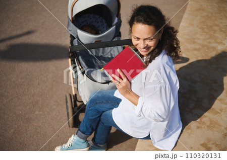 View from above of authentic delightful young mother smiling looking aside, enjoying reading book outdoor View from above of authentic delightful young mother smiling looking aside, enjoying reading book outdoor 110320131