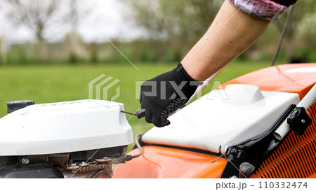 Unrecognisable gardener man in protective gloves starts the lawnmower before cutting green grass lawn in his backyard. Man with motorised lawnmower cares for landscaping lawn 110332474