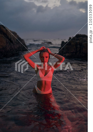 Attractive woman enjoys nocturnal swim in sea illuminated by crimson light. Nighttime ocean dip by stylish female, beauty fashion shoot. Model in swimwear posing, serene water backdrop. Attractive woman enjoys nocturnal swim in sea illuminated by crimson light. Nighttime ocean dip by stylish female, beauty fashion shoot. Model in swimwear posing, serene water backdrop. 110333099