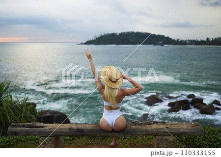 Woman in bikini with a beautiful physique sitting on beach looking at ocean view on sunset in swimwear. Slim figure girl enjoys sea vacation with straw hat in hands. Pretty slender fit blonde female. 110333155