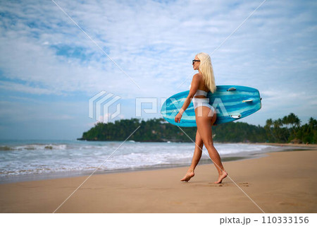 Blonde woman with sunglasses walks on sandy beach carrying blue surfboard, ready to surf waves by tropical coast. Female surfer enjoys summer day at sea shore preparing for water sports. 110333156