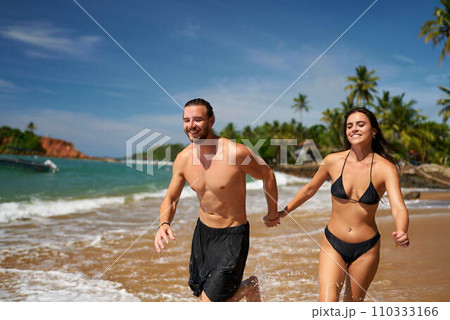 Joyful couple runs on tropical beach, hand-in-hand. Athletic man and slender woman exercise by sea. Fit pair enjoys sunny vacation, laughs during scenic seaside sprint. Outdoor activity, health, fun. 110333166