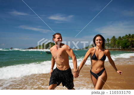Fit couple runs on tropical beach, playful man and woman exercise near ocean. Athletic pair in swimwear enjoys summer vacation, jogging along shoreline, healthy lifestyle, beach workout, sunny day. 110333170