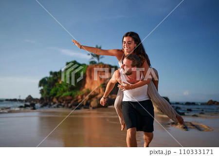 Joyful couple enjoys piggyback ride on sandy beach, woman points ahead, man carries, blissful vacation moment at sunset, casual beachwear, fun dating app concept, active summer leisure, sea backdrop. 110333171