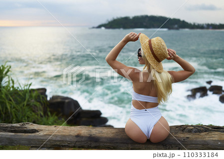 Woman in bikini with a beautiful physique sitting on beach looking at ocean view on sunset in swimwear. Slim figure girl enjoys sea vacation with straw hat in hands. Pretty slender fit blonde female. Woman in bikini with a beautiful physique sitting on beach looking at ocean view on sunset in swimwear. Slim figure girl enjoys sea vacation with straw hat in hands. Pretty slender fit blonde female. 110333184