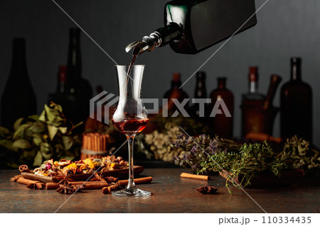 Brown herbal liquor or balm is poured from a vintage bottle into a glass. 110334435