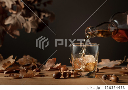 Whiskey with ice on a wooden table with dried-up oak leaves. 110334438