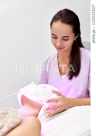 woman receiving led light therapy mask treatment from beautician in beauty salon 110335827