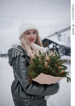 Portrait of young blond woman in beige coat walking in the city with a bouquet of fresh fir branches 110336831