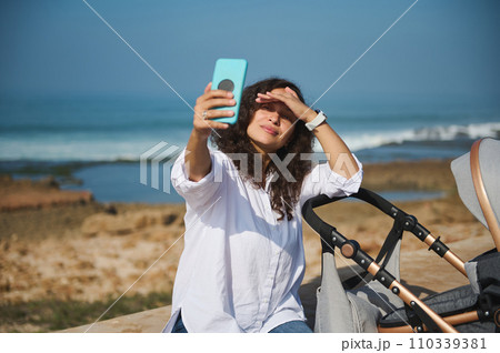 Happy young mom smiles broadly while takes selfie on her modern smart phone, walking with her baby in baby carriage Happy young mom smiles broadly while takes selfie on her modern smart phone, walking with her baby in baby carriage 110339381