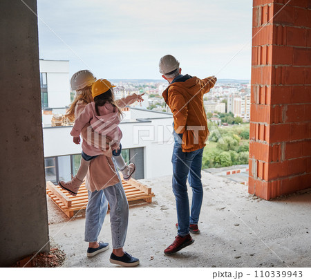 Back view of parents with daughter standing inside apartment building under construction. Man and woman with child wearing building helmets while enjoying city view from future home. 110339943