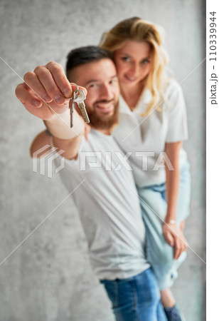 Cheerful man holding wife and smiling while showing apartment keys. Happy couple real estate house buyers demonstrating keys from new home. Focus on male hand with keys. 110339944