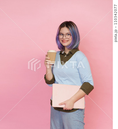 Smiling pupil girl teenager, wearing glasses, immersed in studies with folded laptop and cup of coffee or tea on pink background. Student showcases joyful expression, embodying essence of modern 110342077
