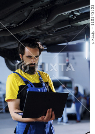 Diligent mechanic in garage using laptop to follow checklist while doing maintenance on car. Meticulous employee in auto repair shop does checkup on vehicle helped by device 110345303