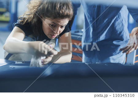 Hardworking repairman in garage finishing mending car, looking underneath vehicle hood to remove remaining oil leaks. Employee does annual engine cleaning on customer automobile in auto repair shop 110345310
