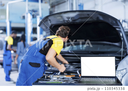Mockup laptop on car service work station bench with technician in blurry background replacing engine. Isolated screen device next to qualified garage expert fixing client automobile Mockup laptop on car service work station bench with technician in blurry background replacing engine. Isolated screen device next to qualified garage expert fixing client automobile 110345338