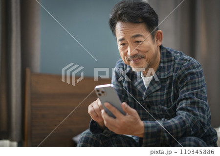 ベッドに座ってスマートフォンの画面を見るパジャマ姿の中年男性 110345886