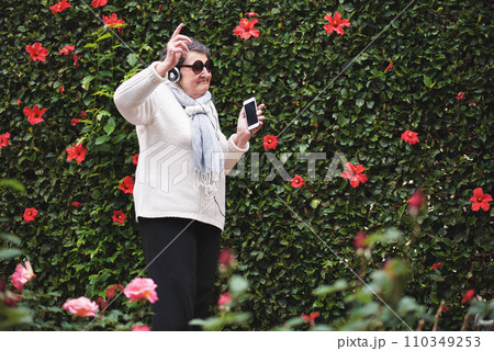 Funny old woman dancing listening to music on smartphone wearing earphones smiling enjoying fun celebrating retirement in garden with flower wall 110349253