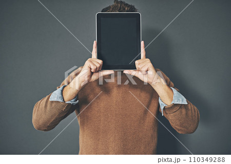 Young man holding a blank tablet up to his face, in casual wear against a gray background with copy space. Alone male standing with a blank screen pressed against his head. Adult posing with a device 110349288