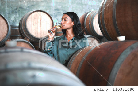 Woman doing wine tasting, drinking glass of chardonnay or sauvignon blanc in winery cellar amongst barrels on vineyard. Beautiful oenologist or sommelier enjoying a relaxing, luxury beverage indoors. 110351090
