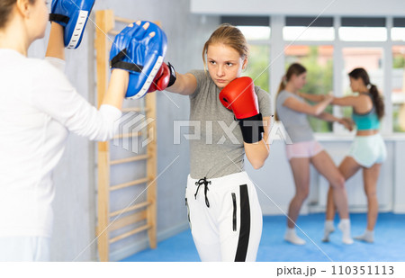 Adolescent girl in boxing gloves punching pads in hands of coach in gym Adolescent girl in boxing gloves punching pads in hands of coach in gym 110351113