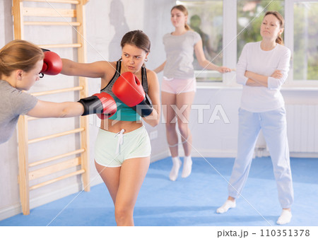 Two teenage girls in sportswear are engaged in boxing sparring in the gym under guidance of trainer in gym Two teenage girls in sportswear are engaged in boxing sparring in the gym under guidance of trainer in gym 110351378