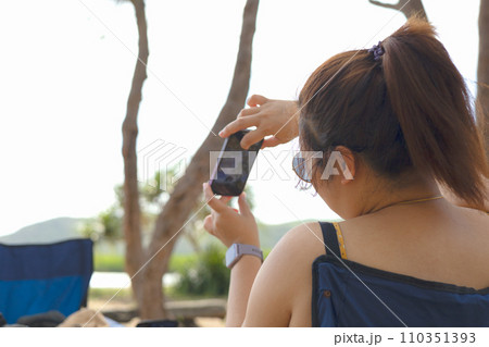 Woman wearing dark glasses smiling and taking a photo with smartphone in the park. 110351393