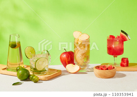 Glasses of lemon, kumquat, apple and watermelon detox drinks are displayed on a white table with a green background. Cooling drink rich in vitamins. 110353941