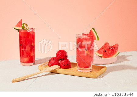 Watermelon is a popular summer cooling fruit. Glasses of juice and fresh watermelon are displayed on a white table with a pastel pink background. Front view. 110353944