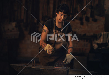Hard work is always the way. a young woman hitting a hot metal rod with a hammer in a foundry. 110355786