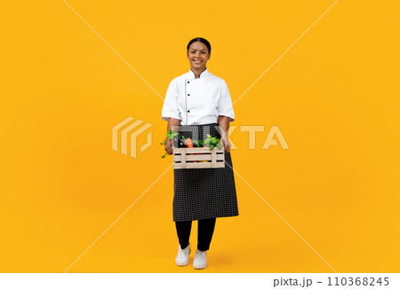 Full-length portrait of black female chef holding crate full of fresh vegetables 110368245