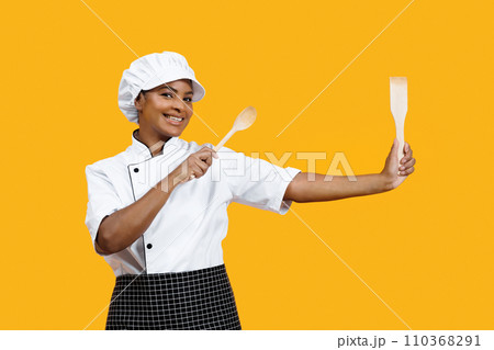Friendly black female chef with radiant smile holding cooking utensils Friendly black female chef with radiant smile holding cooking utensils 110368291