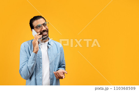 A man with a beard and glasses cheerfully talks on the phone, gesturing with his free hand 110368509