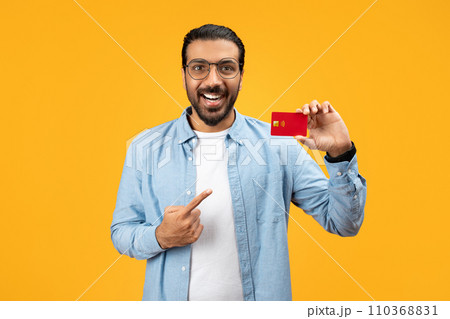 A cheerful man in a denim shirt holding a red credit card and pointing at it with his other hand 110368831