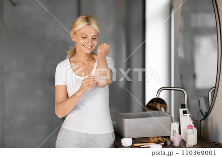 Smiling mature woman in casual clothes applying body lotion to her elbow 110369001