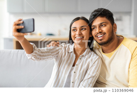 Cheerful young indian couple sitting on sofa and taking selfie on smartphone Cheerful young indian couple sitting on sofa and taking selfie on smartphone 110369046