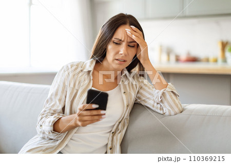 Frustrated young woman looking at smartphone while sitting on couch at home 110369215