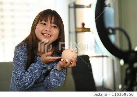 Pretty asian girl holding piggybank in front of camera while recording video on smartphone. Saving money for future concept Pretty asian girl holding piggybank in front of camera while recording video on smartphone. Saving money for future concept 110369356