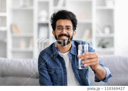 Cheerful young Indian man in denim jacket holding glass of water Cheerful young Indian man in denim jacket holding glass of water 110369427
