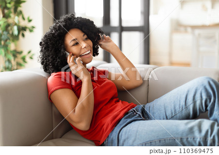Happy black woman relaxing on sofa while having a pleasant phone conversation Happy black woman relaxing on sofa while having a pleasant phone conversation 110369475