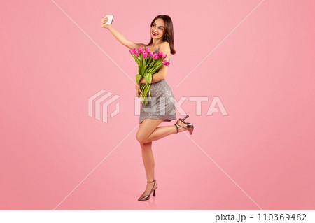 Joyful woman in a shimmering dress capturing a playful moment with a selfie 110369482