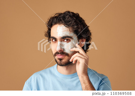 Calm brunette with curly hair, bearded man with dermatology facial care mask and looking at camera against beige studio background. Calm brunette with curly hair, bearded man with dermatology facial care mask and looking at camera against beige studio background. 110369508