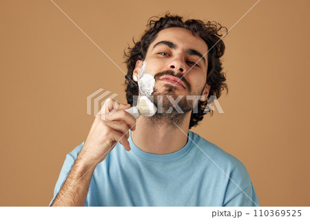 Young brunette man with curly hair applies shaving foam with retro shaving brush against sandy studio background. Young brunette man with curly hair applies shaving foam with retro shaving brush against sandy studio background. 110369525