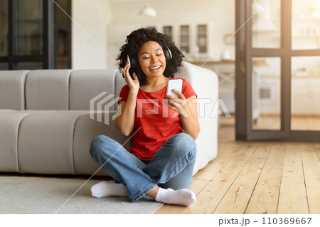 Cheerful young black woman sitting on floor, listening music on her smartphone Cheerful young black woman sitting on floor, listening music on her smartphone 110369667