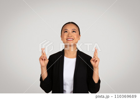 Happy young asian woman in suit crossed fingers, looking up at empty space 110369689